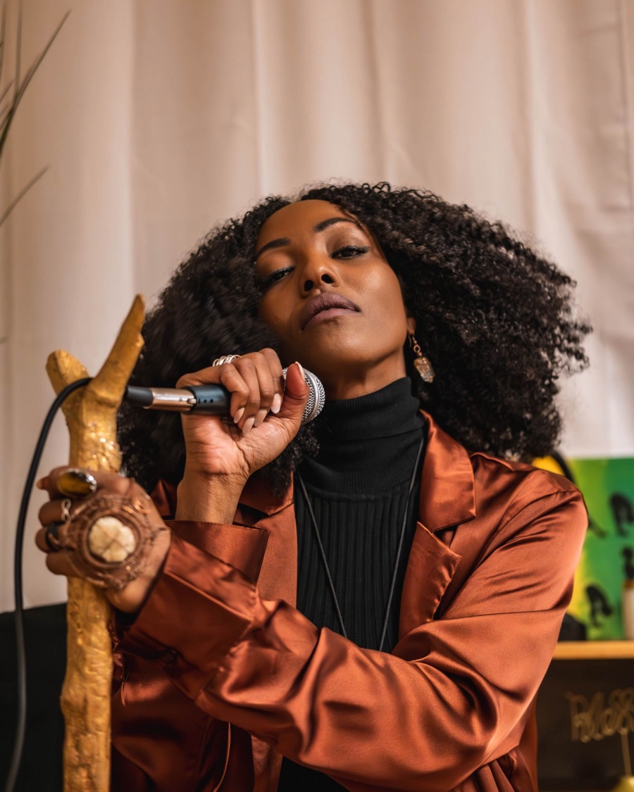 A performer in a copper satin jacket singing into a microphone, holding a gold staff